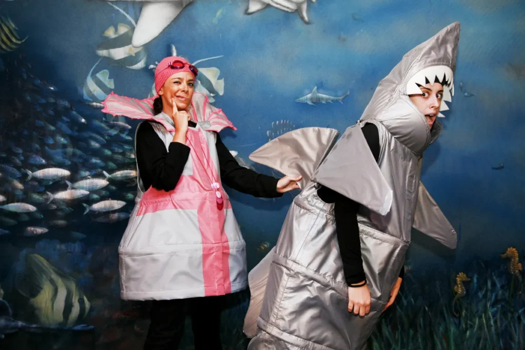 Dos actores disfrazados de criaturas marinas posando frente al fondo submarino durante una actividad educativa teatral.